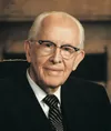 Photograph of President Ezra Taft Benson. He is seated in a leather chair in front of a fireplace. His hands are clasped in front of him and he is wearing a large turquoise ring on one finger. Official portrait. 1986