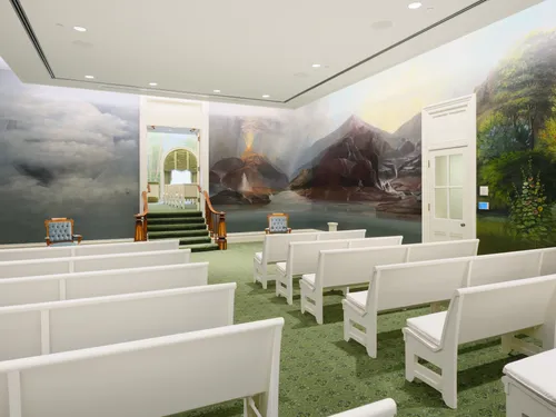 Interior image of the Manti Utah Temple. The image focuses on the Ordinance room. You see pews in the room. Around the walls are murals.