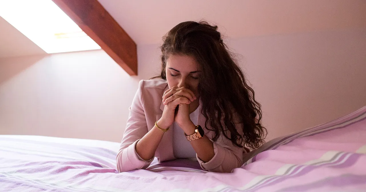 A woman kneels in prayer at her bedside