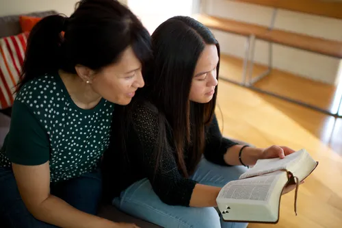 A mother sits down next to her daughter, and they read from the scriptures that the daughter is holding.