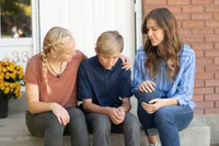 Two young women comforting a young man sitting on his front porch