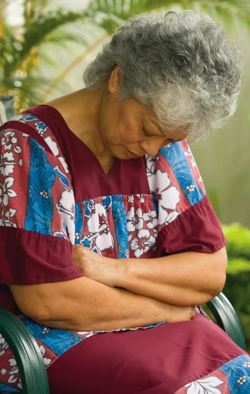 A Fijian woman sitting in a chair.  She has her arms folded and head bowed in prayer.