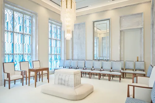Interior image of the Brasilia Brazil Temple Sealing room. The room features an altar in the middle with a chandelier above it. There are chairs on three sides of the room and one wall has stained glass. 