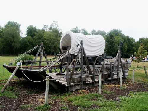 A wooden pioneer wagon with a white canopy sitting on a ferry on display in Nauvoo, Illinois.