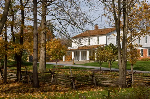 The white clapboard of Newel K. Whitney’s store is viewed through trees with a split-rail fence in the foreground.