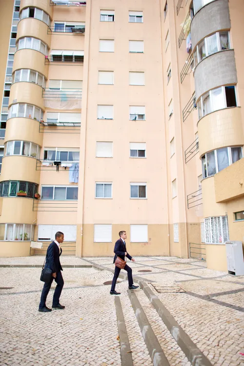 Two elder missionaries in suits, carrying shoulder bags, walk beside tall, cream-colored buildings in Portugal.