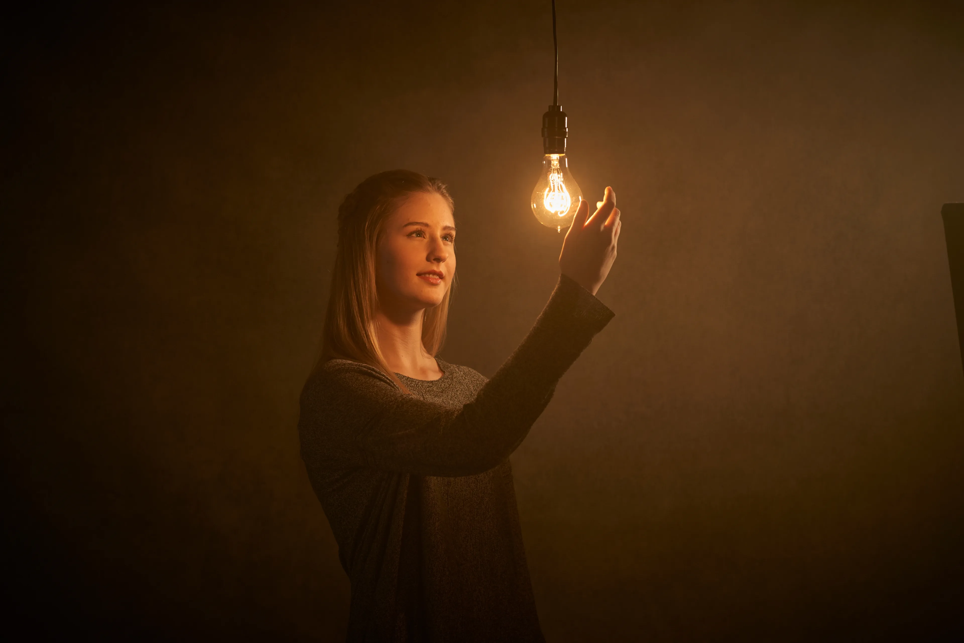 A young woman in a dark room reaching out toward an illuminated light bulb.