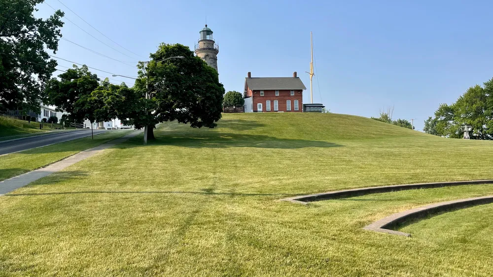 A lighthouse and two-story brick building stand atop a grassy hill.