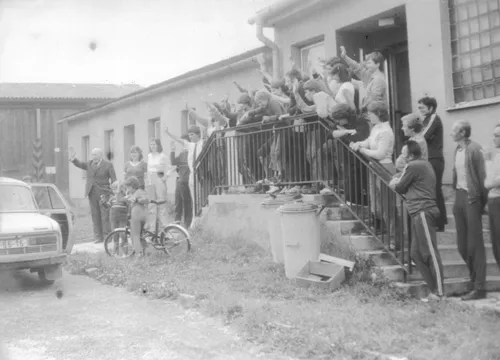 About fifty people stand on the steps of a building and wave to a car.