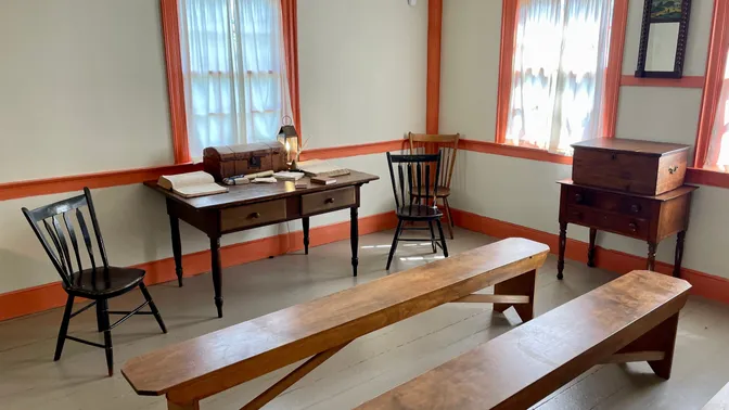 A sparsely furnished room. A desk with books, papers, and a chest stands beneath a window flanked by  chairs. Three benches sit in front of the desk to provide seating observers.