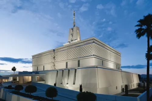 The exterior of the Mexico City Mexico Temple is lit up, viewed from the front entrance side in the evening, with a surrounding landscape of trees.