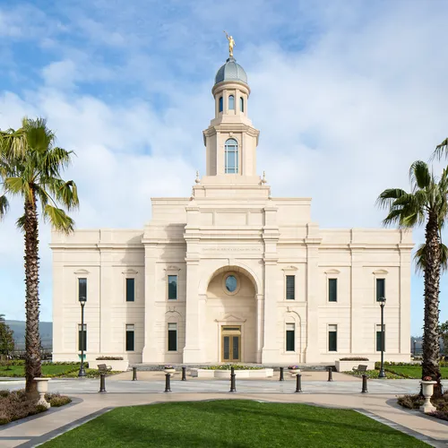 Concepción Chile Temple Exterior. In this image we see the outside of the temple. There are palm trees on the sides. The temple is made of white stone with a blueish domed top. Angel Moroni sits on top. There is a Chilean flag to the side.