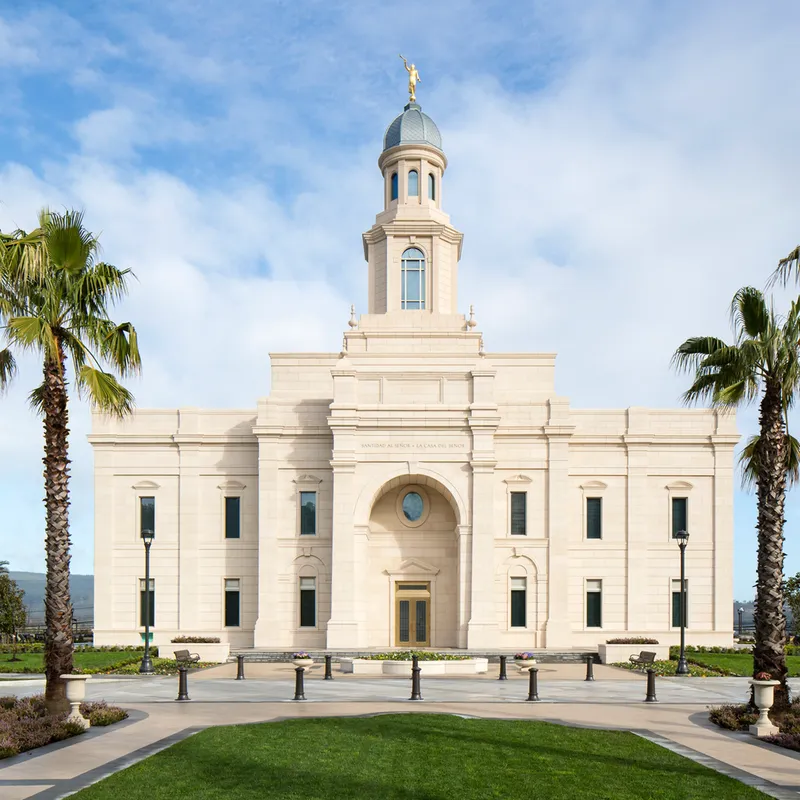 Concepción Chile Temple Exterior. In this image we see the outside of the temple. There are palm trees on the sides. The temple is made of white stone with a blueish domed top. Angel Moroni sits on top. There is a Chilean flag to the side.