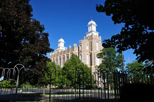The trees on the grounds of the Logan Utah Temple framing a view of the temple, with a deep blue sky in the background and a black gate in the foreground.