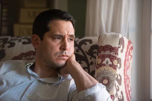 A man sits in a chair.  He looks like he is deep in thought and doesn't like what he is thinking or feeling.  Very sad looking.