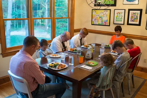 A father, mother, three sons, and a daughter sitting at a table and praying with two elder missionaries before eating their meal.