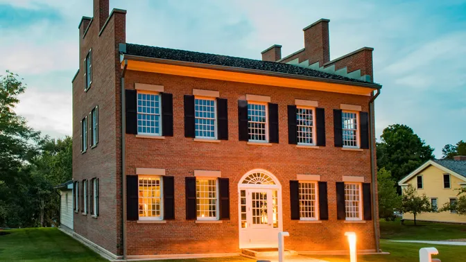 A two story red brick home with many windows. It is illuminated in the twlight by lights on a pathway leading up to the entrance.