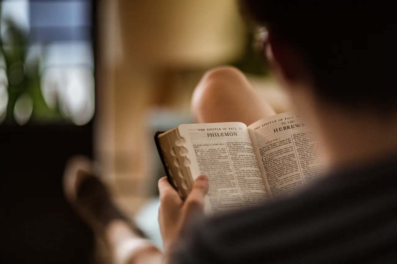 A man sitting on a couch studies the Holy Bible with the pages open to the epistle of Paul to Philemon