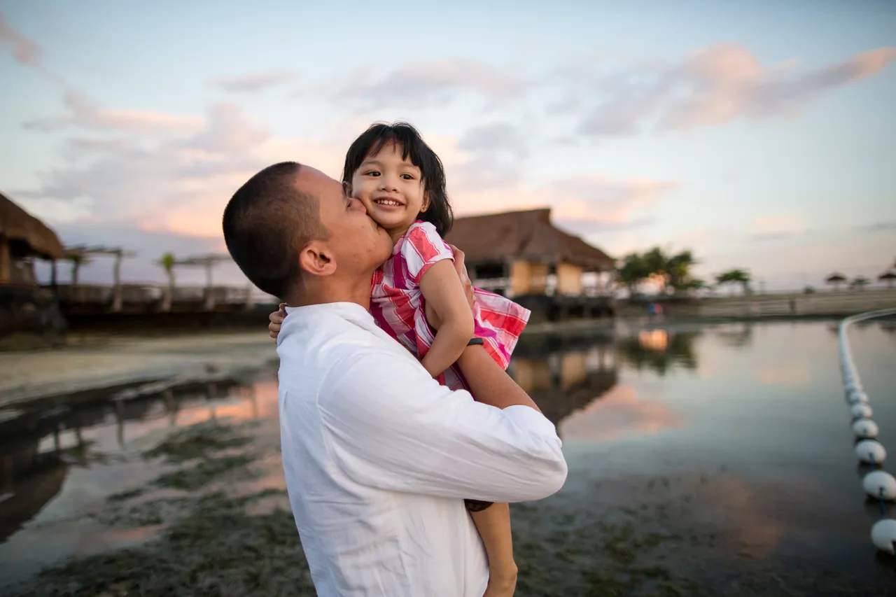 A father picks his young daughter up and kisses her on the cheek