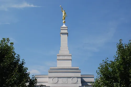 A view of the Palmyra New York Temple spire on a sunny day, with the leaves of two green trees seen toward the bottom of the frame.