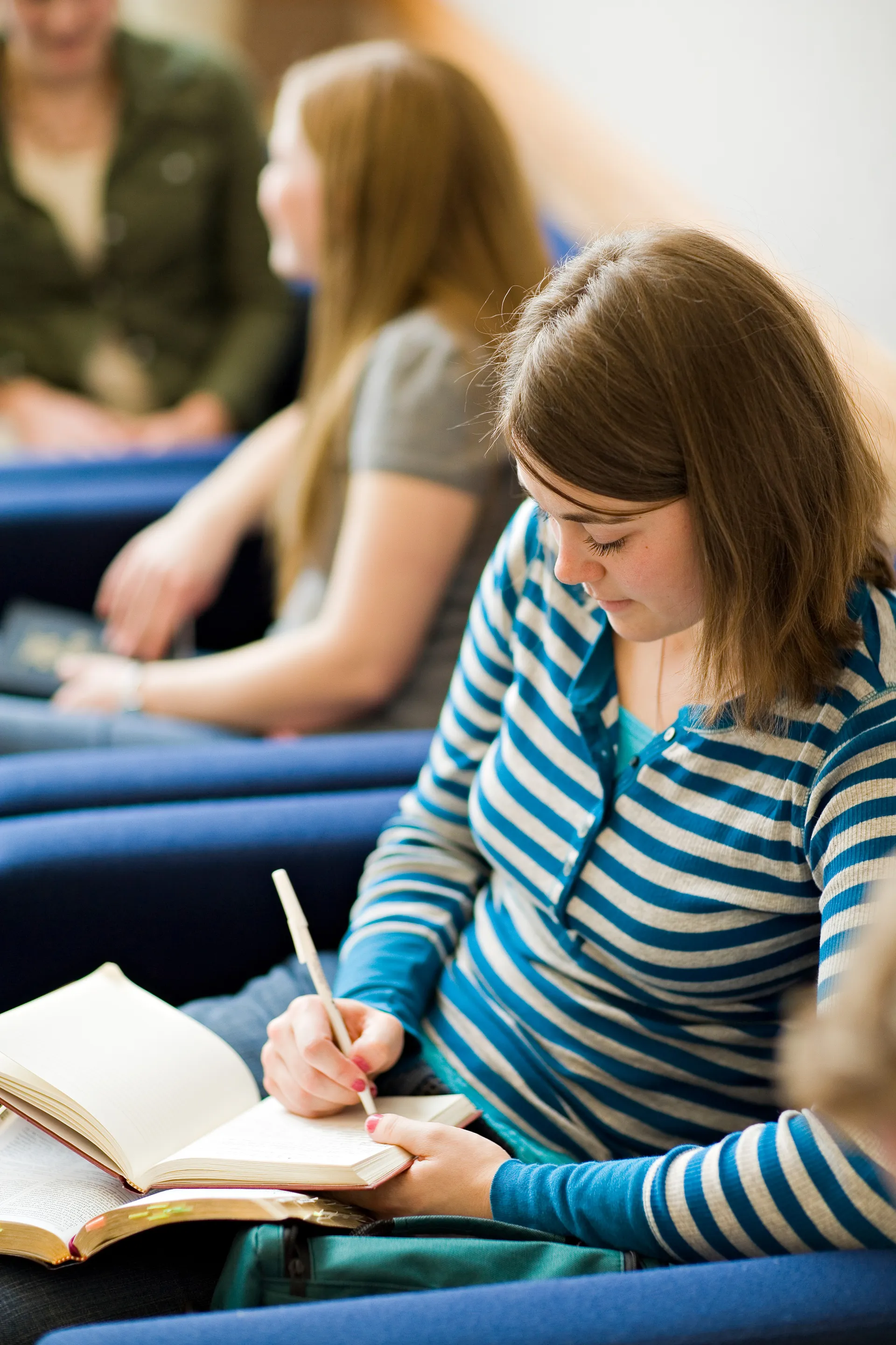 A young woman sits in a chair and writes in a journal while studying scriptures.