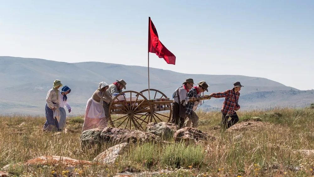 Seven people in pioneer clothing push and pull a handcart over rocky terrain.