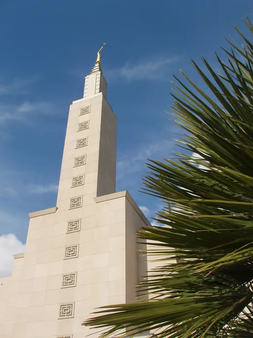 The spire on the Los Angeles California Temple, seen behind a green bush on the temple grounds, with a blue sky overhead.