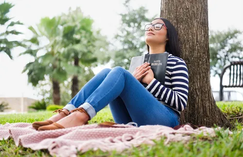 young woman sitting next to a tree and holding a Bible