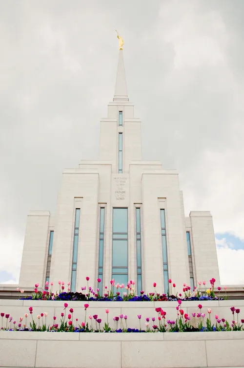 A front view of the Oquirrh Mountain Utah Temple on a spring day, with colored flowers in the front and a dull gray sky above.