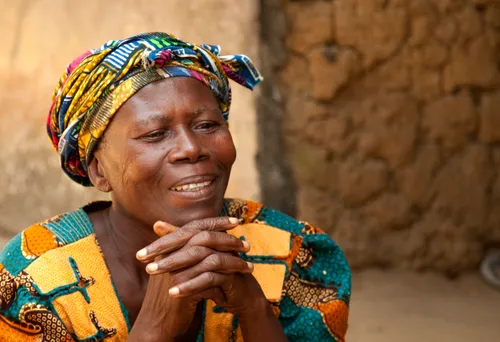 An African woman in a bright blue, yellow, and pink head wrap and shirt, with her chin resting on her two clasped hands.