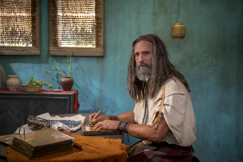Nephi sits at a table with the brass plates and metal plates as he records his testimony.