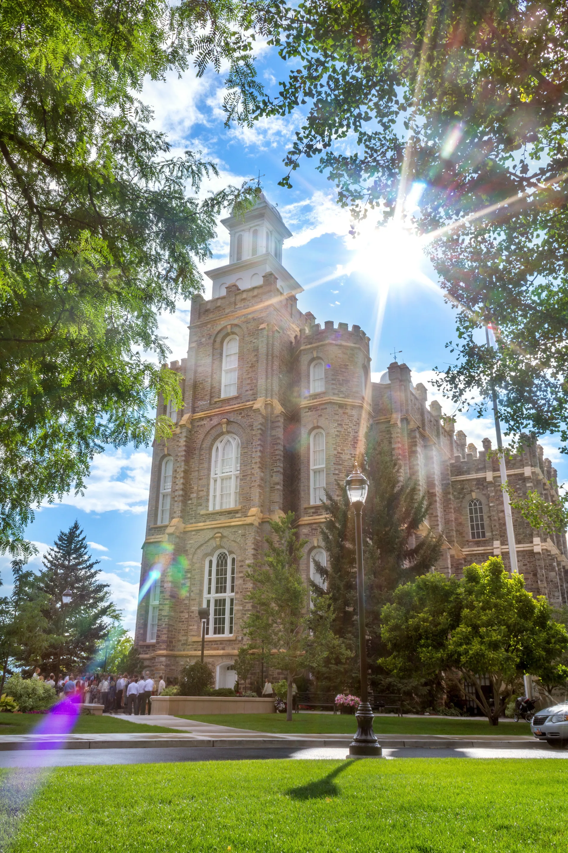 The sun shining above the Logan Utah Temple with a group of people below.