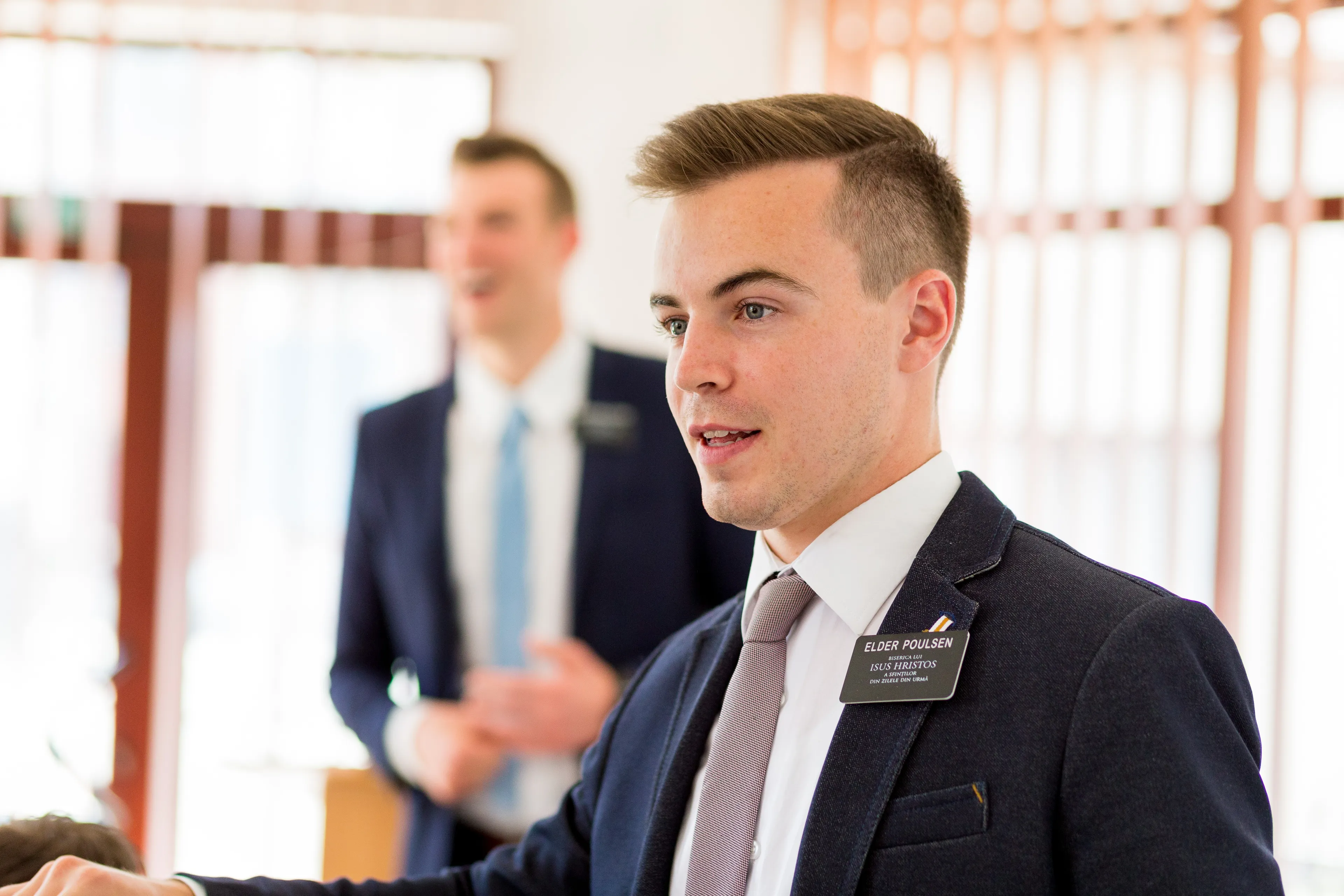 An elder missionary standing in a classroom with his companion in the background.