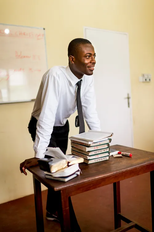 A male Sunday School teacher stands at the front of a classroom and leans his hands on a table where hymnbooks and his scriptures are stacked.