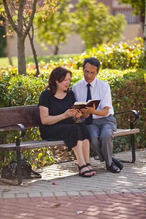 A man in a white shirt, black tie, and gray pants sits on a wooden bench next to a woman in a black dress and sandals as they read the scriptures together.