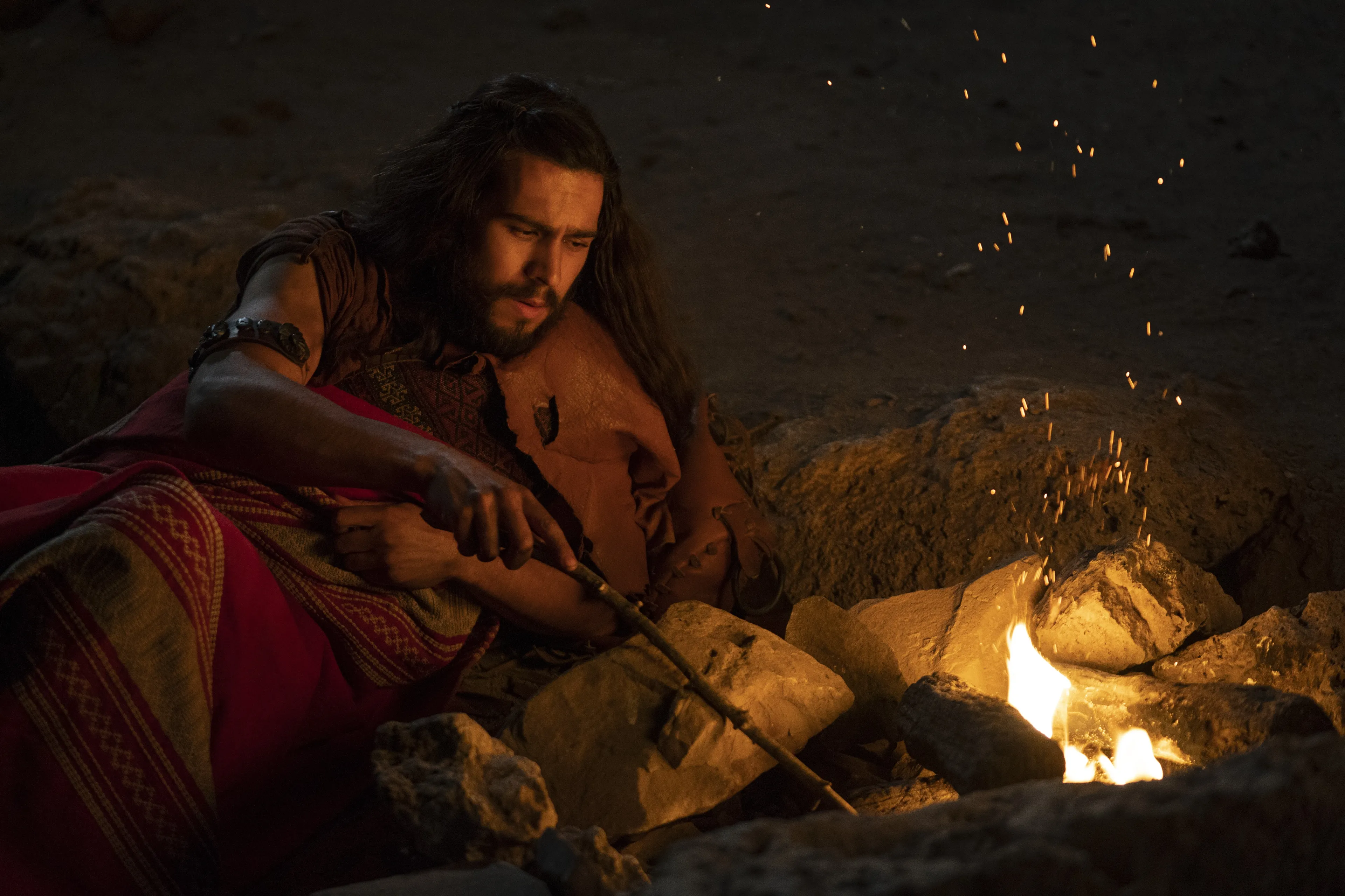 Enos lays next to a fire at night as he is hunting in the wilderness.