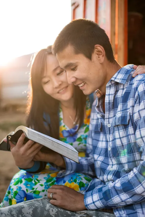 A young man and woman sit side by side outdoors and read from a set of scriptures together.