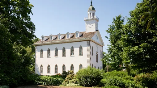 A three-story white stucco building, with dormer windows running the length of the roof shingled. A white tower, topped by a weather vane, stands on the front of the building.