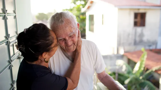An elderly man stands outside in Brazil while his wife kisses his cheek.
