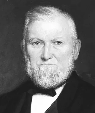 Portrait of Wilford Woodruff. He is depicted wearing a black suit, vest and bow tie, seated in a red chair and resting one arm on the arm rest and the other on a table.