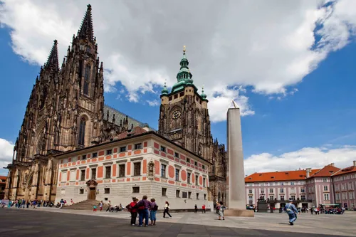 People walking outside the Saint Vitus Cathedral in the Czech Republic with a blue sky and clouds overhead.