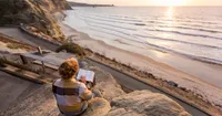 A young adult male sits on a rock at the edge of a cliff. He is reading the scriptures as the sun begins to set.