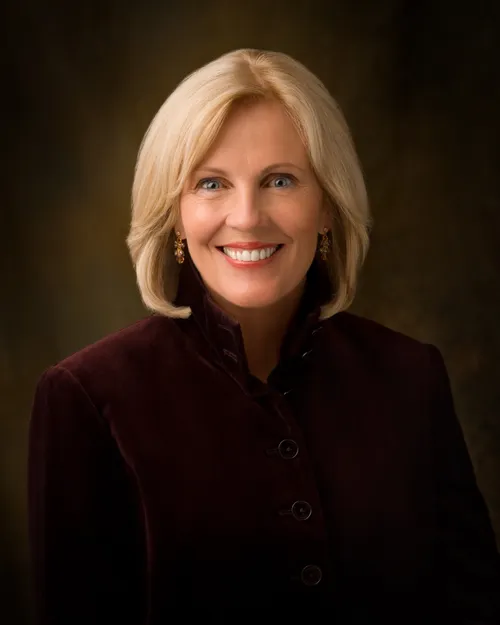 A photograph of Elaine Schwartz Dalton against a brown background, wearing a dark burgundy blazer with buttons down the front.