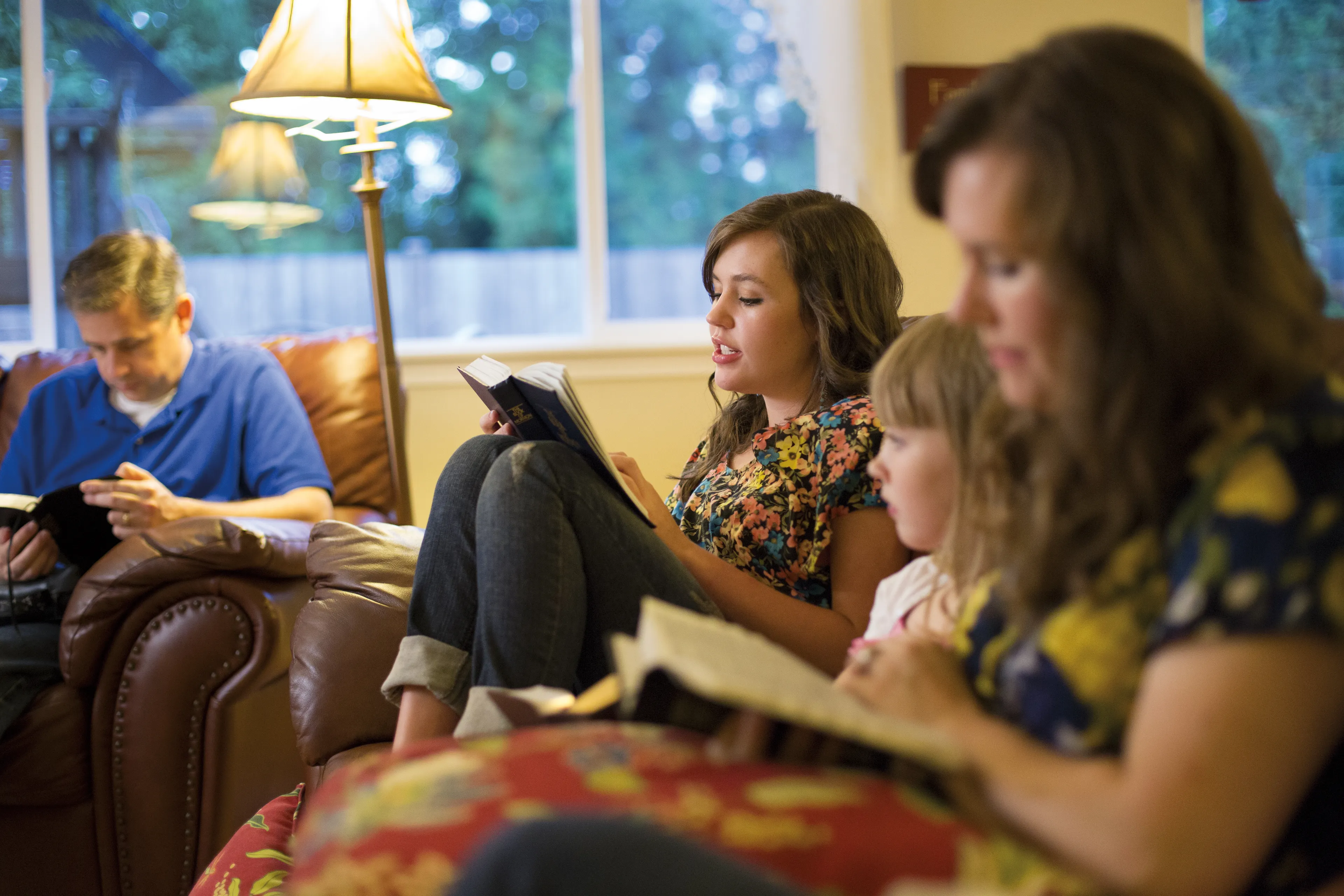 A young woman reads from the scriptures while family members follow along.