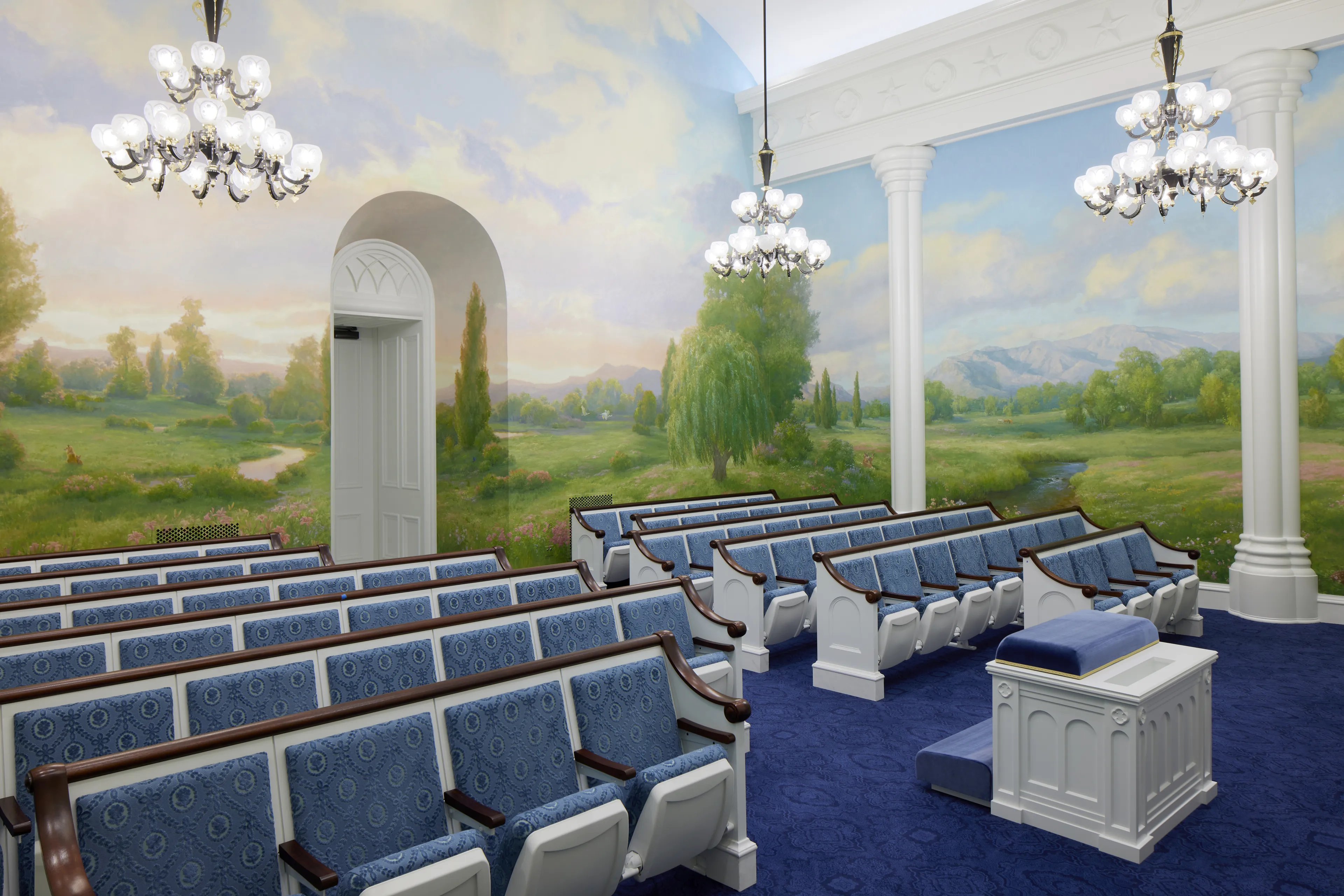Interior image of the St. George Utah Temple. The image features the Instruction Room (Ordinance Room). It features an altar in the front, murals of the area, and chairs. 