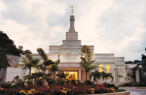 Palm trees, colorful flowers, and a sidewalk in front of the Caracas Venezuela Temple on a cloudy day.