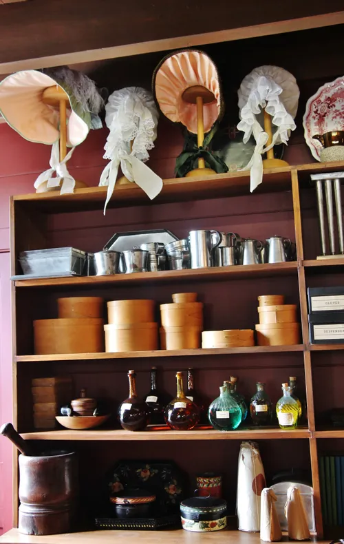 Shelves stacked with goods for purchase, such as round boxes, tin cups, glass bottles, and bonnets with ribbons.