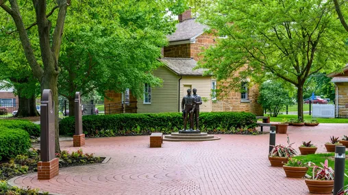 Exterior shot of Carthage Jail, bronze statue of Joseph and Hyrum Smith, and red brick pathway with two stone markers on the left-hand side.