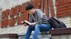 a young man sits on a bench outside and reads the Book of Mormon