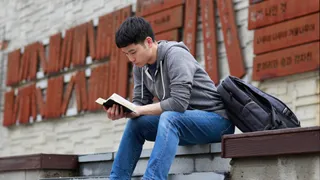 A Korean young man sits on a bench outside and reads the Book of Mormon.